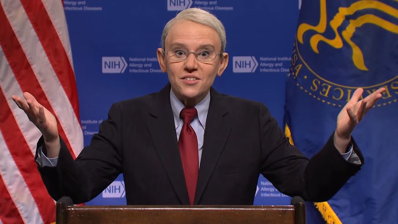 Kate McKinnon as Anthony Fauci, shrugging, at an NIH podium in front of an American flag