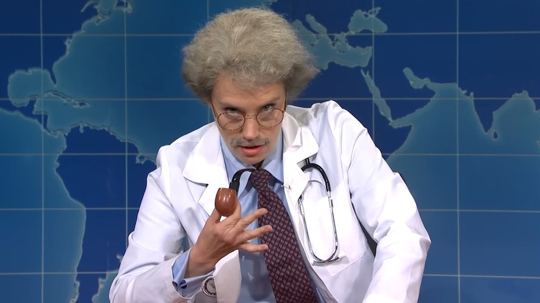 Kate McKinnon in a doctor's coat, holding a pipe, with frizzy grey hair as Dr. Wayne Wenowdis