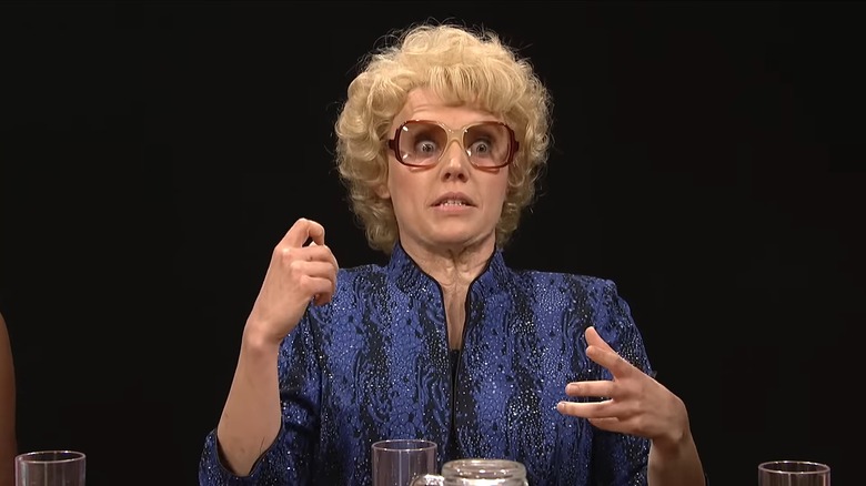 Kate McKinnon with big glasses and a blond wig as Debette Goldry