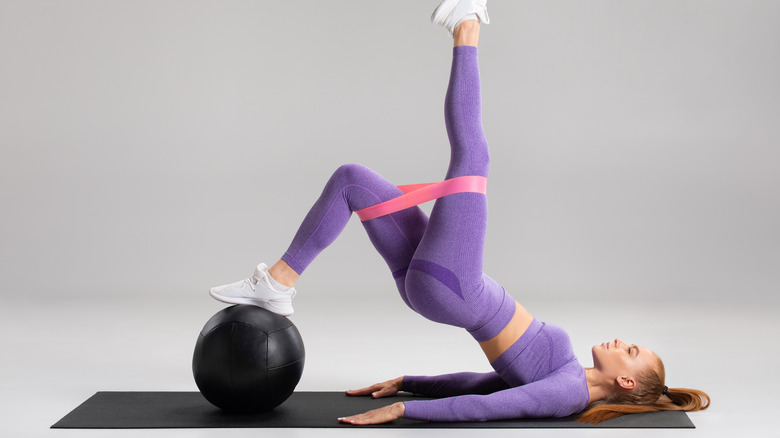 white woman in purple leggings doing single leg glute bridge with resistance band balancing on stability ball