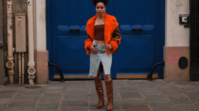 woman in orange coat, denim skirt, and brown boots posing