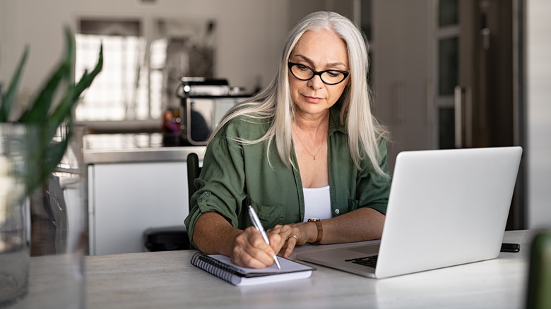 Senior stylish woman taking notes in notebook while using laptop at home.