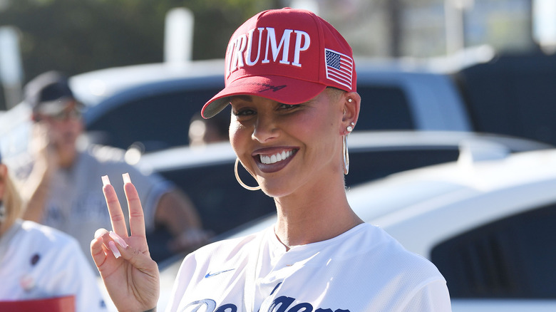 Amber Rose smiles and holds up the peace sign while wearing a red Trump cap