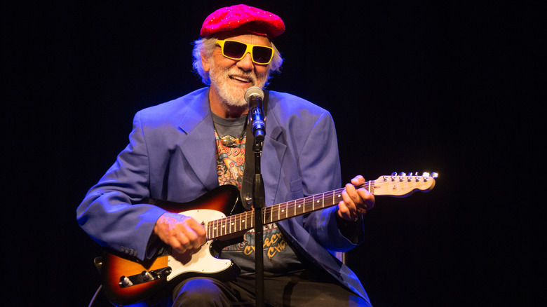 Tommy Chong wearing a hat and sunglasses and playing guitar