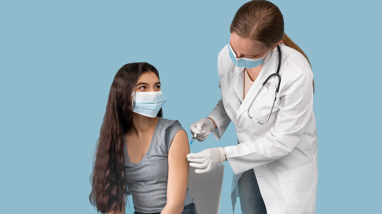 Brunette teen girl wearing a mask and receiving a shot from a masked doctor.