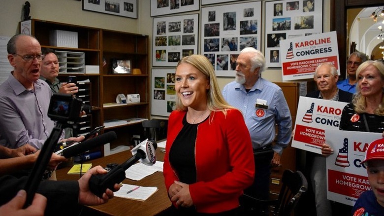 Karoline Leavitt smiling in red jacket and black shirt during interview with supporters holding signs behind her