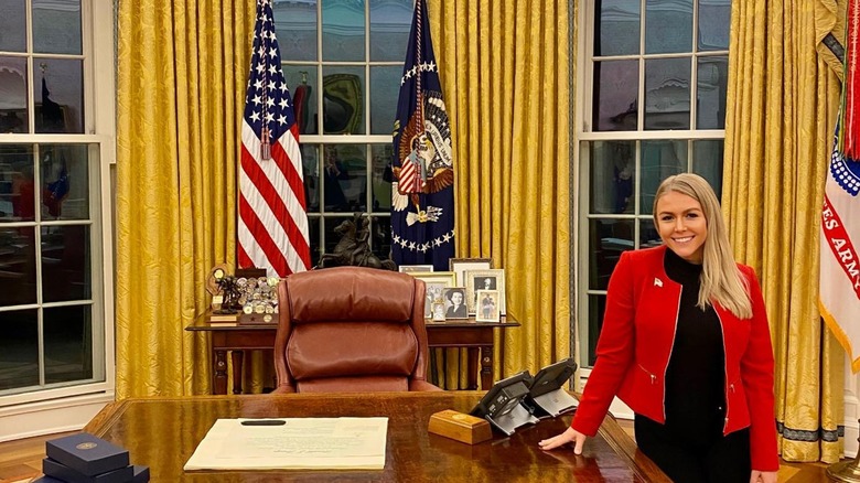 Karoline Leavitt in red jacket and black shirt smiling beside the Resolute desk