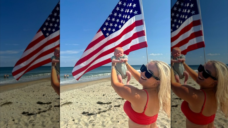 Karoline Leavitt smiling in red bikini on the beach with her baby in front of American flag