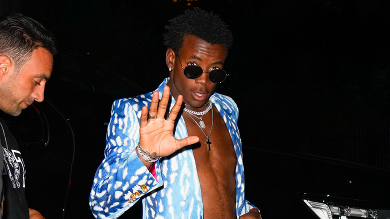 David Banda wearing an opened shirt and sunglasses holding up palm to the camera