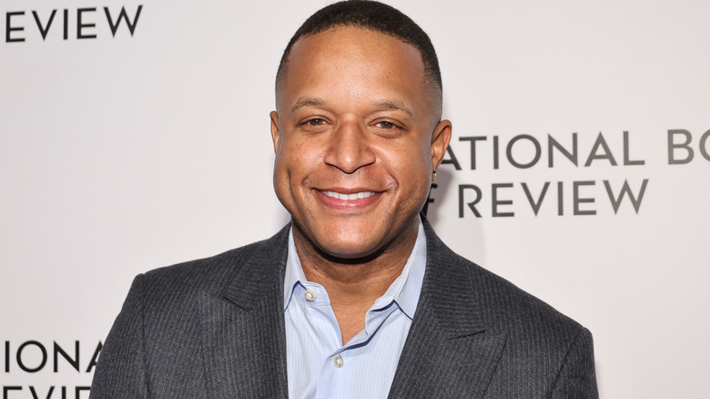 Closeup of Craig Melvin smiling wearing a light blue shirt and gray suit jacket on step-and-repeat