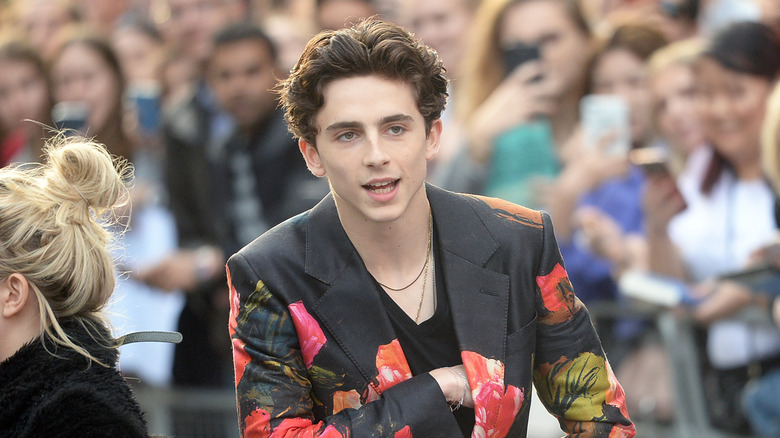 Timothee Chalamet reaching into a floral-patterned suit jacket, fans behind him