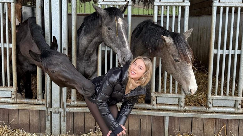 Hannah Selleck smiles crouching down in the middle of three horses at a stable