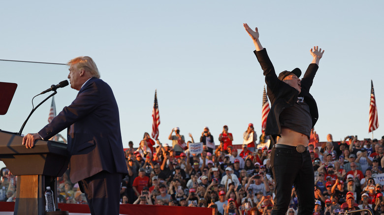 Elon Musk jumping while Donald Trump speaks on a stage