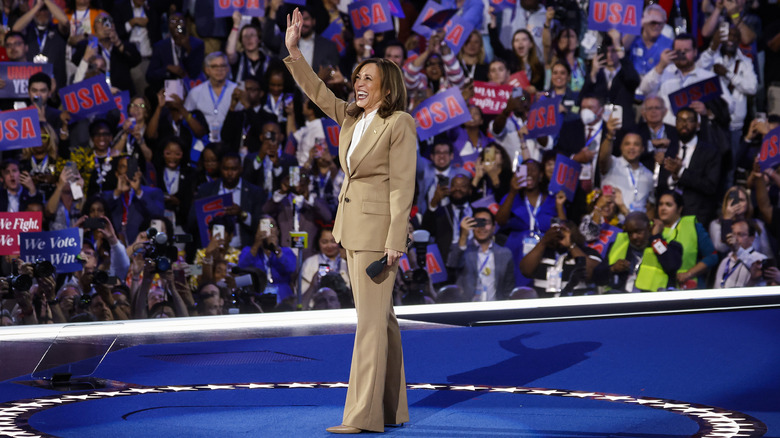 Kamala Harris waving and smiling on stage surrounded by crowds holding USA banners