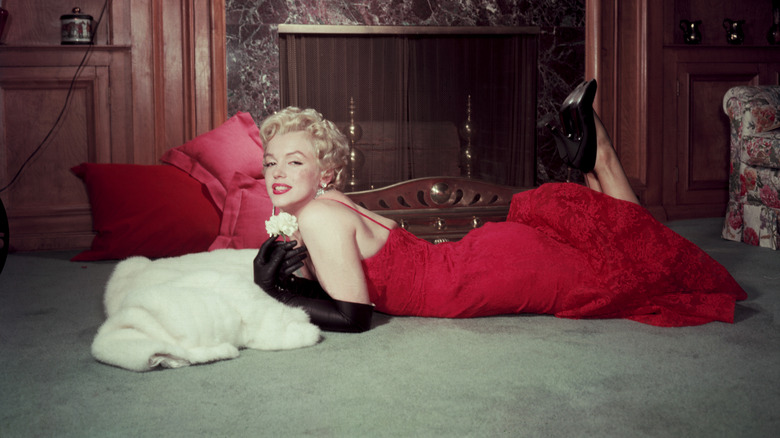 Marilyn Monroe lying on her stomach, wearing a red evening dress, long black gloves, and black pumps poses for a photo