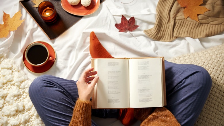 A person reading a book surrounded by coffee, a blanket, and fall leaves