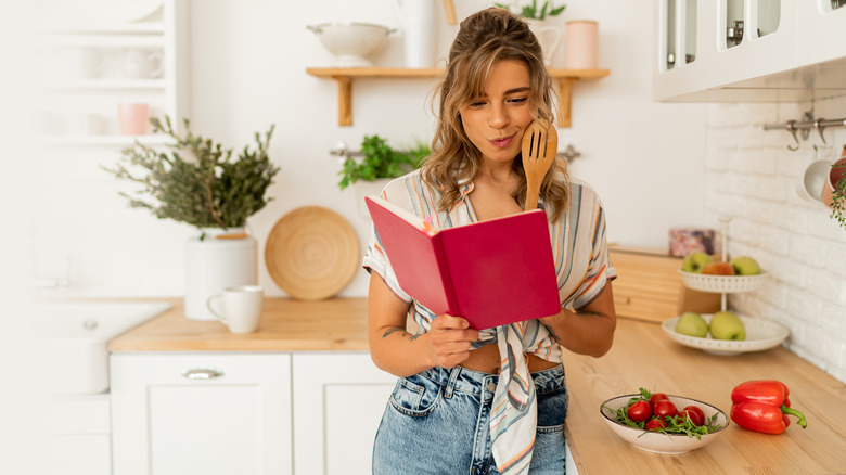 Woman holding a recipe book and wooden spoon while standing in the kitchen