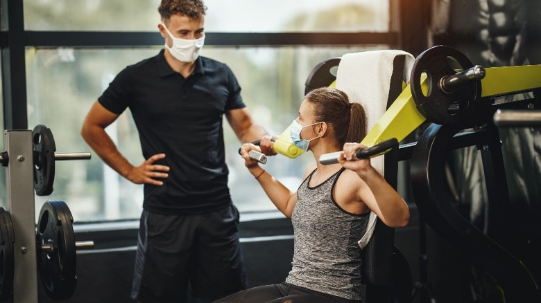 woman using weight machine with trainer at the gym while wearing face mask