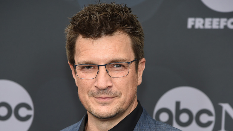 Nathan Fillion smiling while wearing glasses at a press event