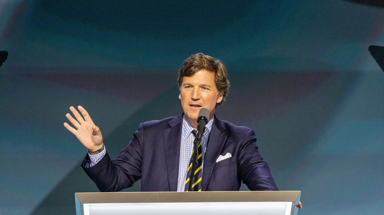 Tucker Carlson stands with his hand raised in front of a podium