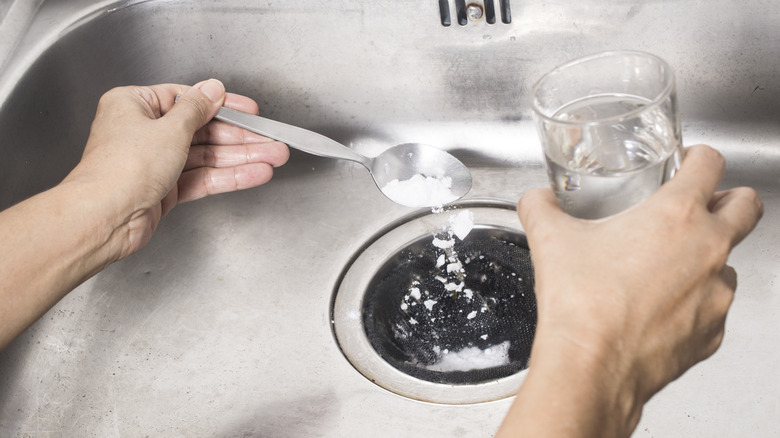 Person putting spoonful of baking soda into a sink drain while holding a glass of vinegar in the other hand