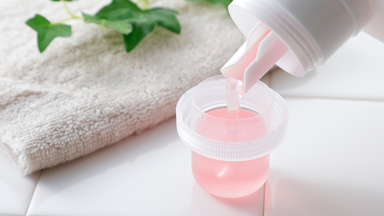 Person measuring pink laundry detergent into a cup sitting on white tile with a towel behind it.. 