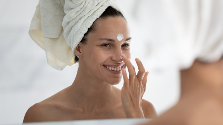 Woman with hair wrapped in towel looks in mirror and applies face cream to forehead and nose while smiling