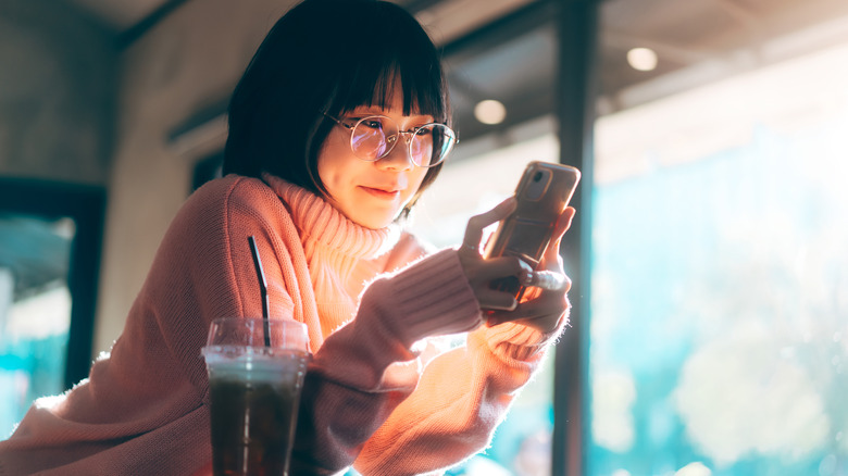 Woman leaning on table in cafe with iced coffee smiling at her phone