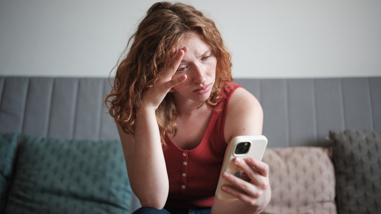 Woman sitting on gray couch with her head in her hand looking at her phone with a pained expression on her face