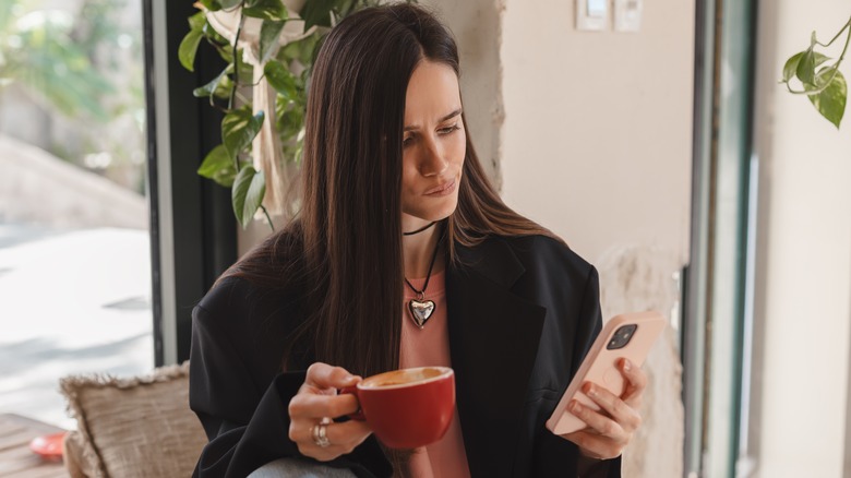 Woman holding cappuccino with a skeptical expression looking at her phone