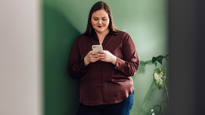 Woman standing against a green wall and green plant smiling as she looks at her phone