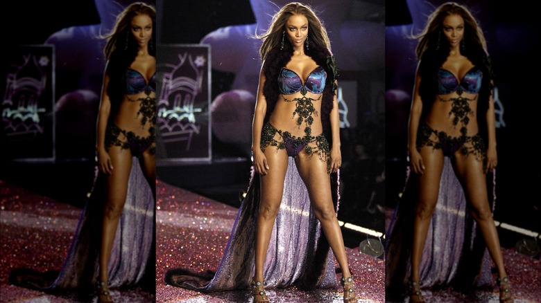 Tyra Banks on Victoria's Secret runway wearing blue jeweled panties and satin bra with dangling jewels and a cape.