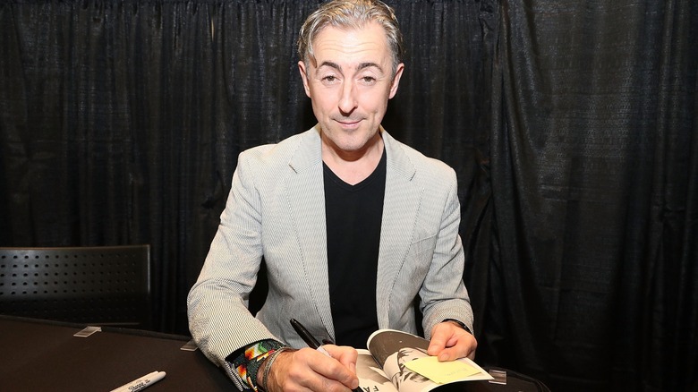 Alan Cumming in a grey suit, signing a copy of his memoir