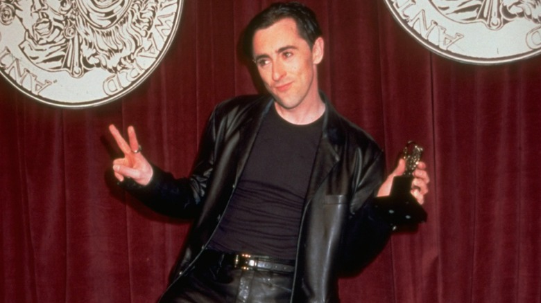 Alan Cumming in a leather jacket, holding his Tony Award, giving a peace sign in 1998