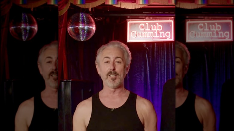 Alan Cumming in a tank top standing in front of a disco ball and the Club Cumming sign