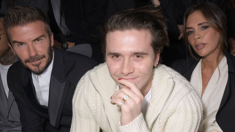 Brooklyn Beckham wearing red nail polish and sitting next to his parents David and Victoria Beckham