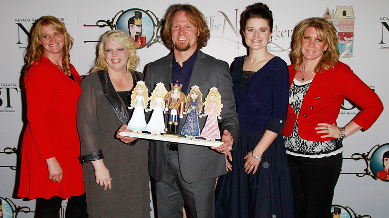 Kody Brown posing with wives on step-and-repeat holding arrangement of paper dolls on either side of a king figurine