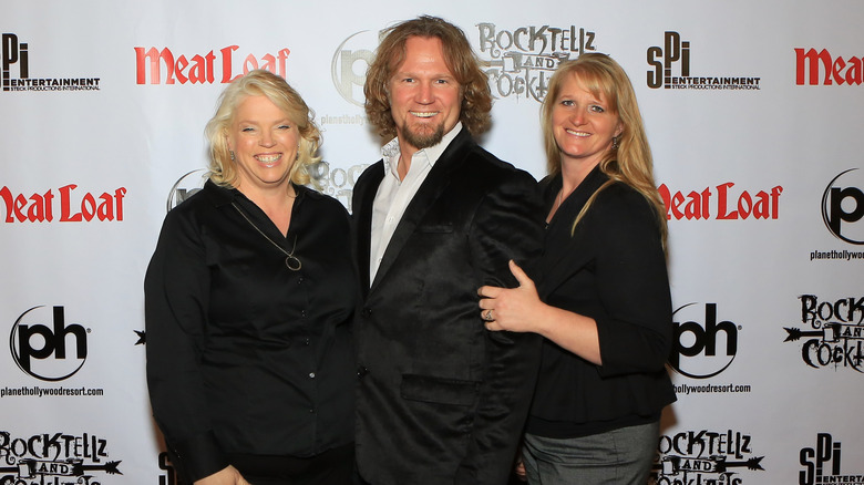 Kody Brown posing with wives Janelle and Christine on step-and-repeat