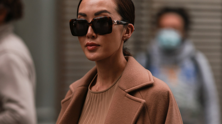 model wearing brown knit top, brown trench coat, and tortoiseshell sunglasses on a busy city street 
