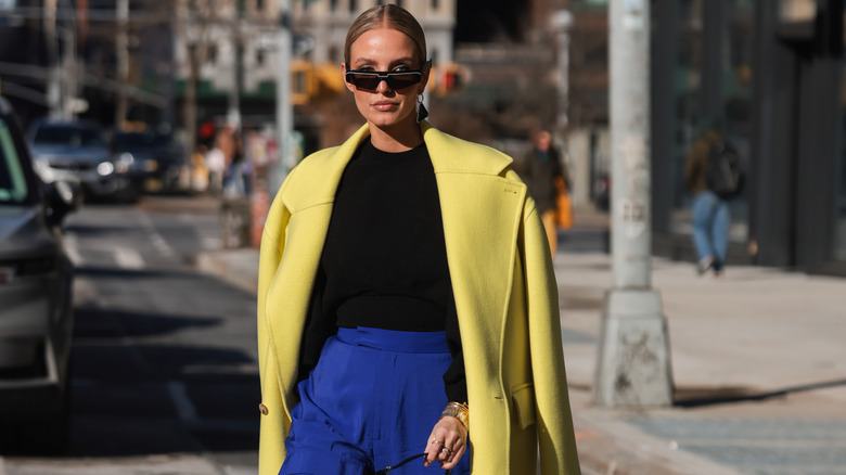 model wearing cobalt blue pants, yellow trench coat, wearing dark sunglasses, walking through a busy street