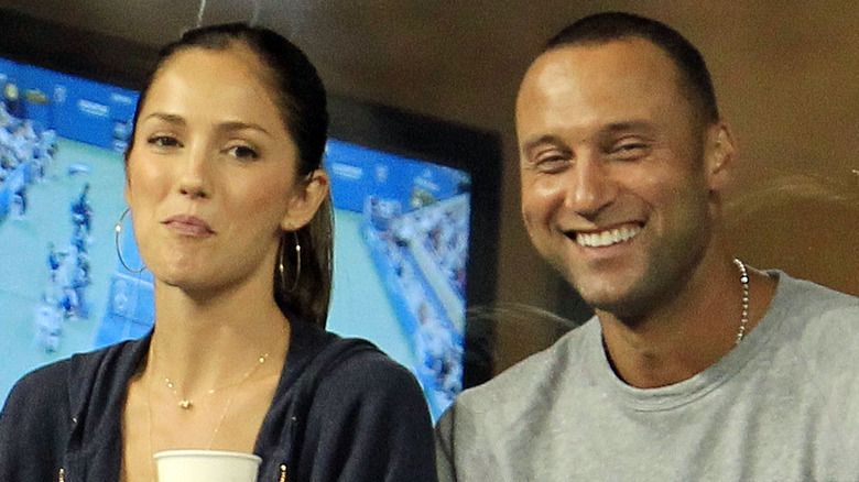 Minka Kelly smiles with hoop earrings as Derek Jeter laughs in a gray shirt and necklace.