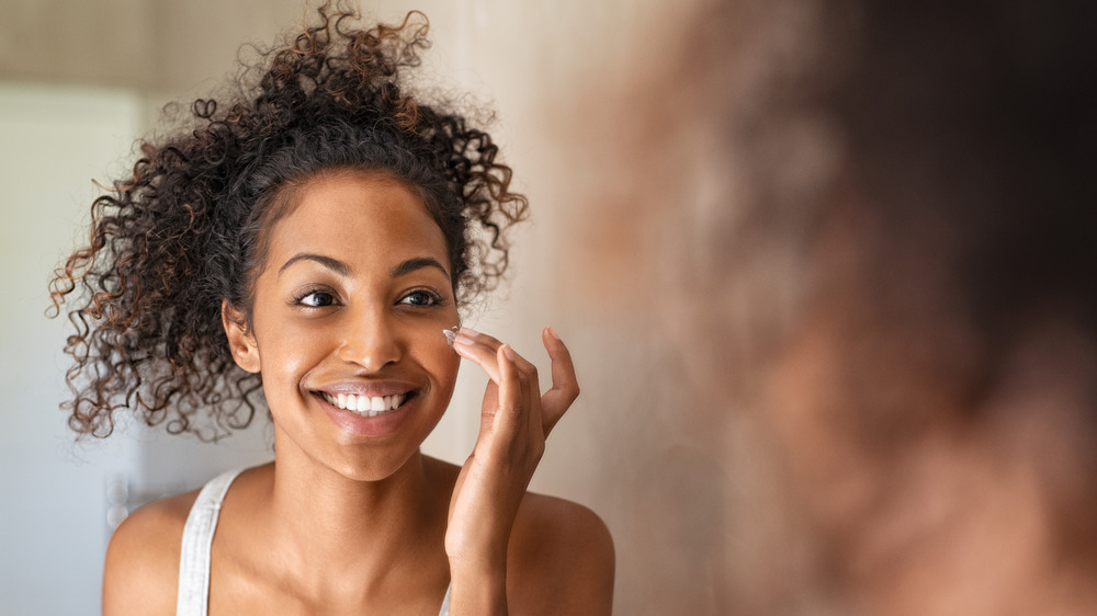 Curly haired woman smiling while applying face cream 