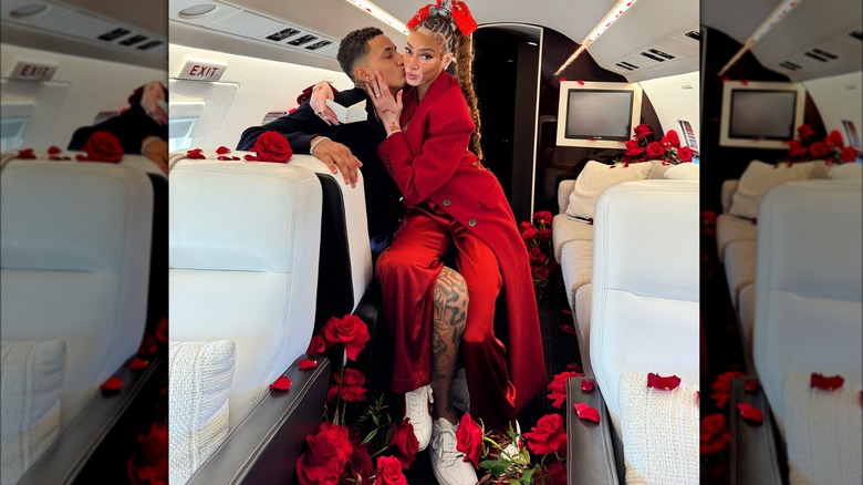 Winnie Harlow showing off her engagement ring while sitting on Kyle Kuzma's lap aboard a small airplane, filled with red roses. Kuzma is kissing her cheek.