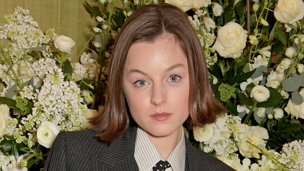 Emma Corrin wearing a suit in front of flowers