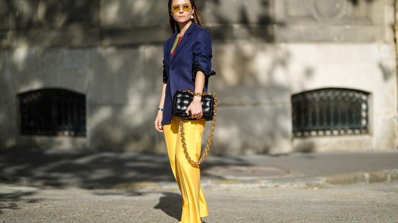 Julia Comil wears yellow sunglasses, gold pearls earrings, a green / blue / brown / red striped pullover, a navy blue long blazer jacket, gold rings, yellow flared flowing pants, a black shiny braided leather handbag from Bottega Veneta.