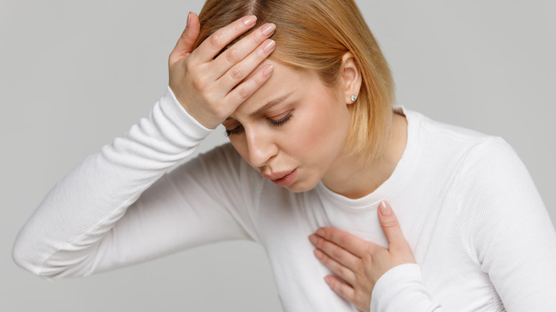 Woman with short blond hair and white longsleeved shirt looking down with one hand on forehead and one hand on chest, blowing air out of her mouth like she is having trouble breathing