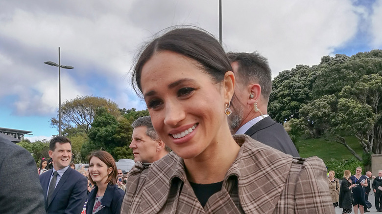 Meghan Markle smiling outside in a crowd wearing a plaid jacket