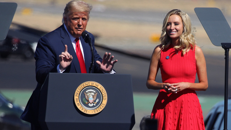 Kayleigh McEnany smiling behind Donald Trump as he delivers a speech