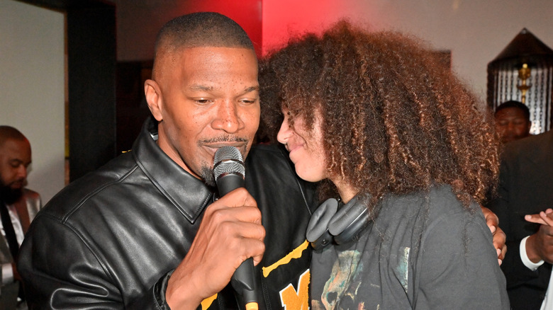 Jamie Foxx speaking into a microphone next to his daughter