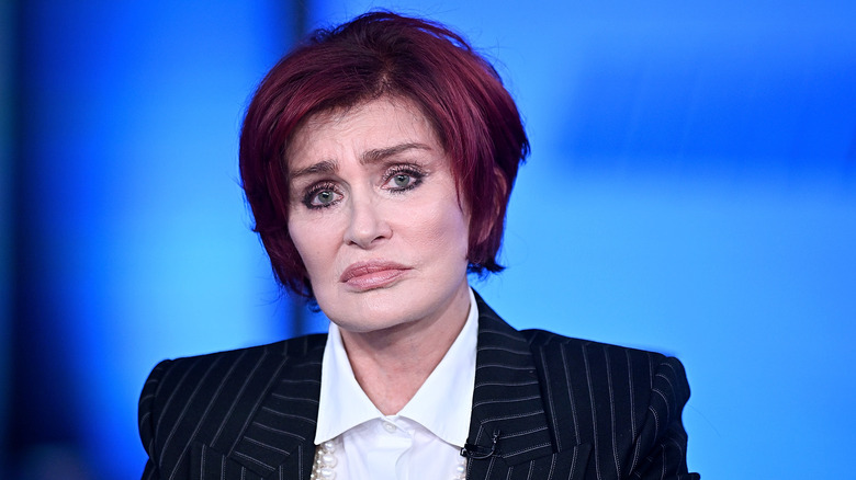 Sharon Osbourne frowning in a suit against a blue background